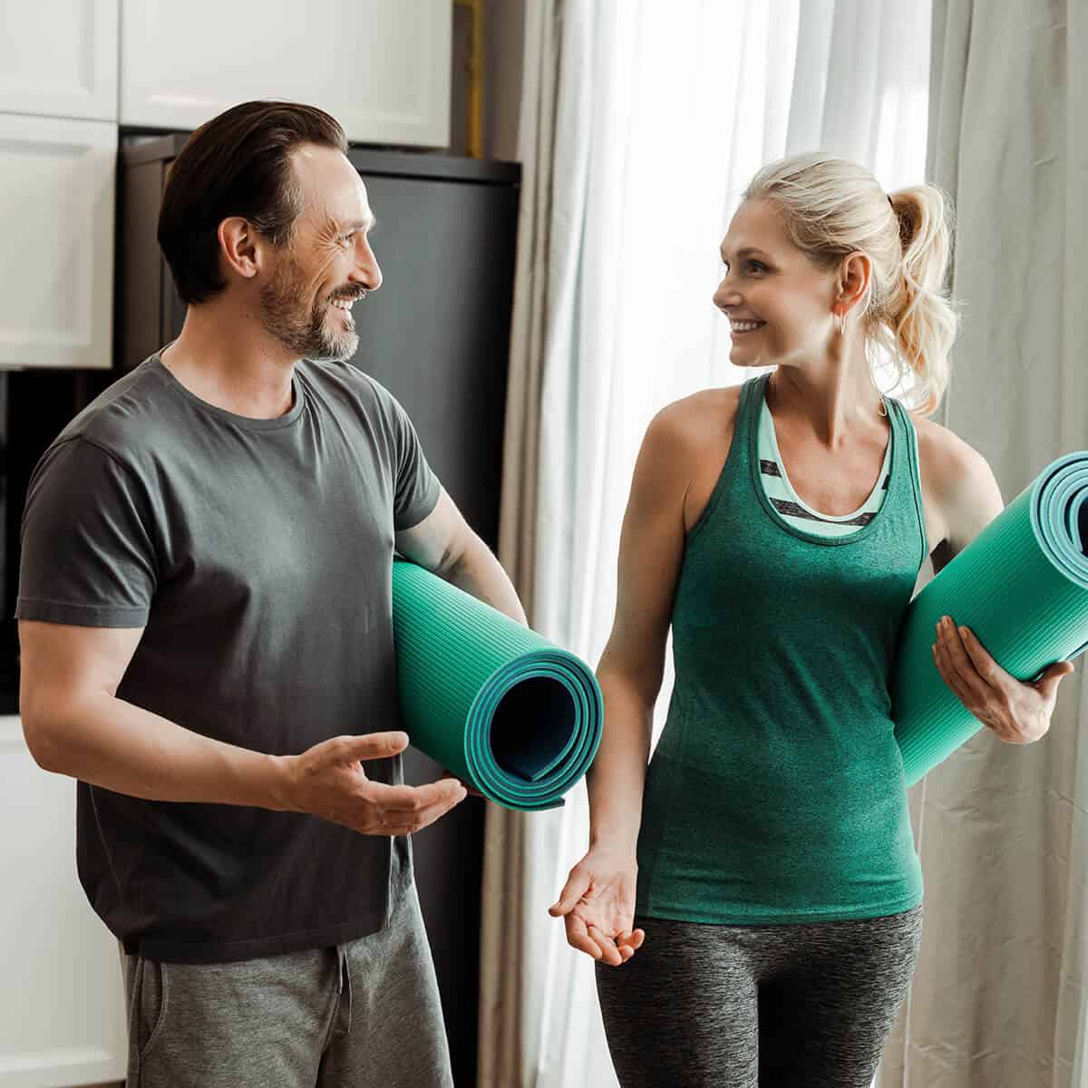 Mature couple in fitness clothes holding exercise mats and smiling at each other
