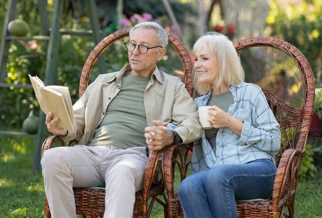 Loving elderly couple reading book together, sitting in wicker chairs and spending spring evening in their garden